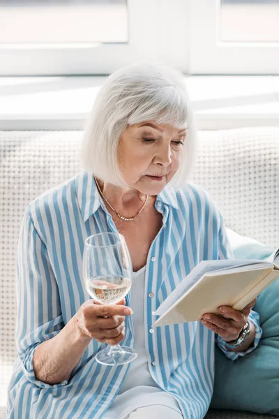 Portrait of focused senior woman with glass of wine reading book on couch at home — Stock Photo