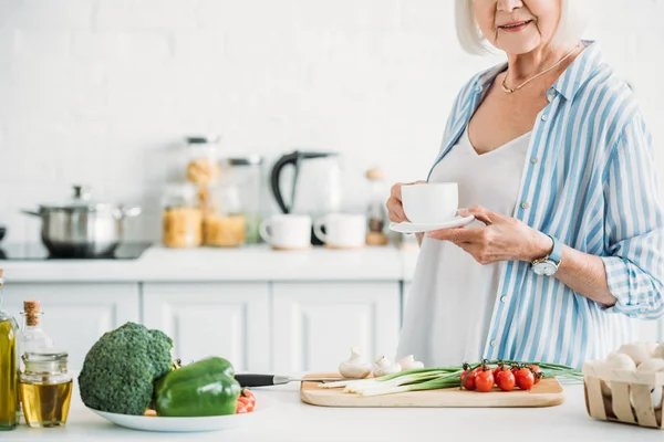 Tiro recortado de mujer mayor con taza de café de pie en el mostrador con verduras frescas para la cena en la cocina - foto de stock