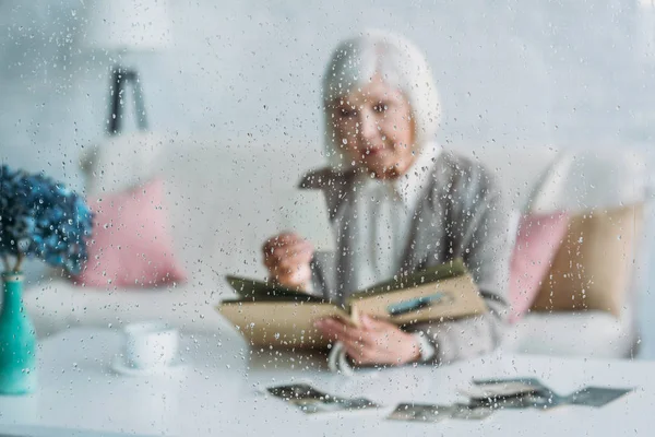 Selective focus of senior woman looking at photographs in photo album at table with cup of coffee at home — Stock Photo