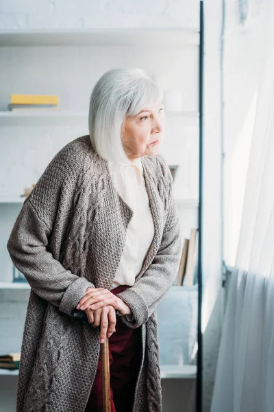 Seitenansicht der nachdenklichen Seniorin mit Gehstock, die zu Hause im Zimmer steht — Stockfoto