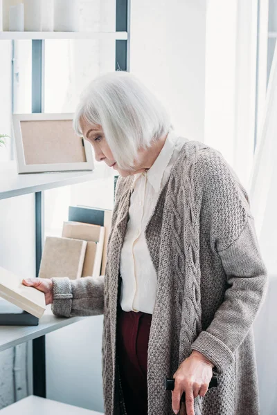Vista lateral da mulher idosa com bengala escolhendo livro para ler em casa — Fotografia de Stock