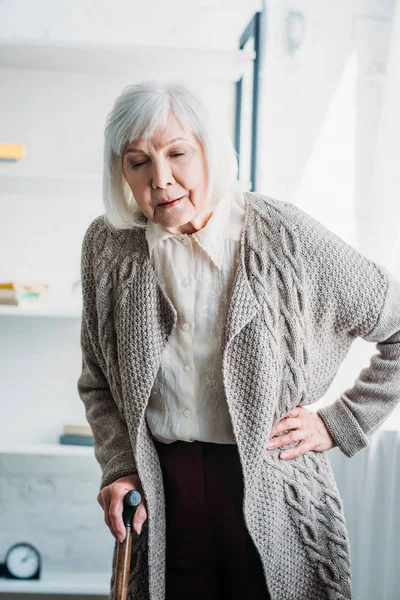 Portrait de dame aux cheveux gris akimbo avec bâton de marche en bois debout au milieu de la pièce à la maison — Photo de stock