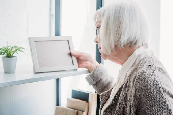 Side view of senior lady looking at empty photo frame on bookshelf at home — Stock Photo