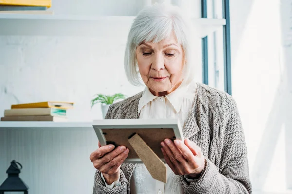 Portrait of thoughtful senior lady in knitted jacket looking at photo frame in hands at home — Stock Photo