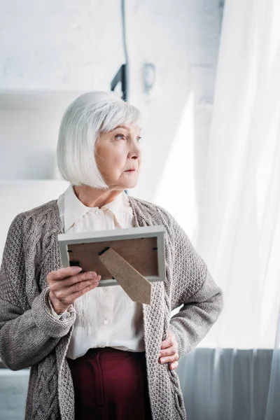 Portrait of pensive senior lady akimbo with photo frame in hand looking away at home — Stock Photo