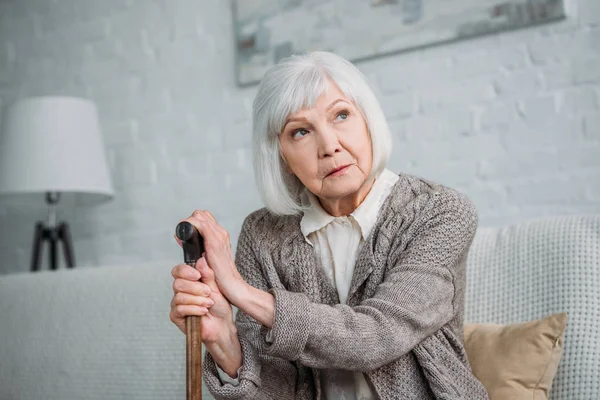 Portrait of pensive grey hair lady with wooden walking stick looking away while resting on sofa at home — Stock Photo