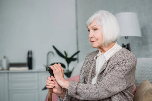 Vista laterale della signora capelli grigi con bastone da passeggio in legno appoggiato sul divano di casa — Foto stock