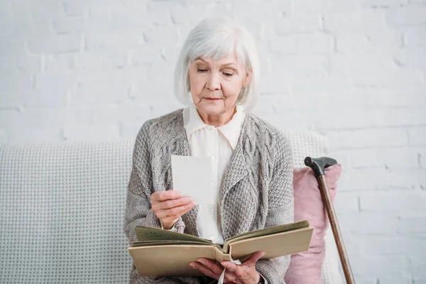 Retrato de la señora de pelo gris mirando fotos del álbum de fotos mientras descansa en el sofá en casa — Stock Photo