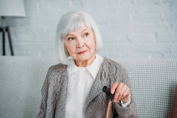Retrato de señora de pelo gris reflexivo con bastón de caminar descansando en el sofá en casa - foto de stock