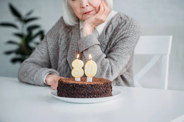 Vista parcial da senhora sênior sentado à mesa com bolo de aniversário com velas sozinho em casa — Fotografia de Stock