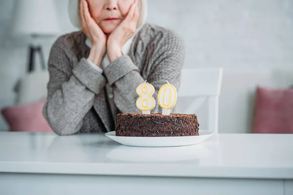 Tiro recortado de senhora sênior sentado à mesa com bolo de aniversário com velas sozinho em casa — Fotografia de Stock