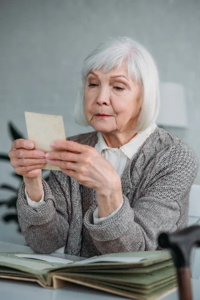 Retrato de mujer de pelo gris mirando la foto del álbum de fotos en la mesa en casa - foto de stock