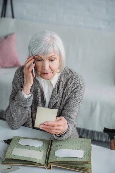 Porträt einer Seniorin mit Foto in der Hand, die zu Hause mit dem Smartphone spricht — Stockfoto