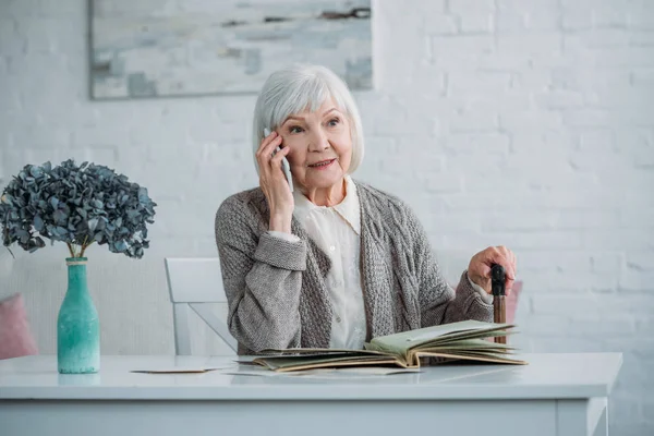 Portrait of senior woman with walking stick talking on smartphone while sitting at table with photo album at home — Stock Photo