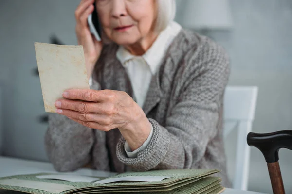 Частичный вид пожилой женщины с фотографией в руке, разговаривающей на смартфоне дома — стоковое фото