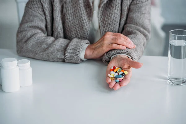 Tiro cortado de mulher idosa com pílulas na mão sentado à mesa sozinho em casa — Fotografia de Stock
