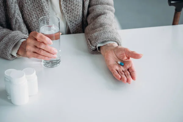 Частичный вид пожилой женщины с таблетками и стаканом воды в руках, сидящей за столом в одиночестве дома — стоковое фото
