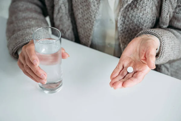 Частковий вид на старшу жінку з таблеткою і склянкою води в руках, сидячи за столом наодинці вдома — стокове фото