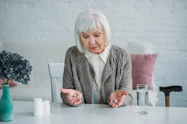 Retrato de mujer de pelo gris con pastillas en las manos sentada a la mesa sola en casa - foto de stock