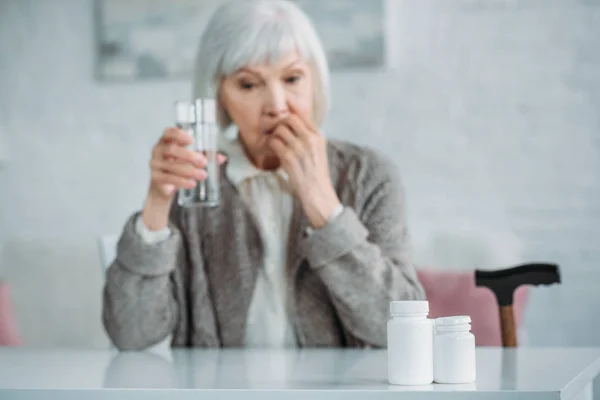 Selektiver Fokus der grauhaarigen Frau mit einem Glas Wasser, das Medikamente zu Hause einnimmt — Stockfoto