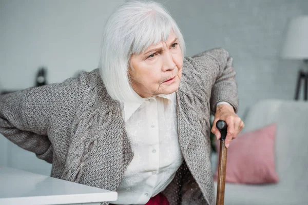 Retrato de senhora de cabelo cinza com dor nas costas e bengala de madeira sentada na cadeira em casa — Fotografia de Stock