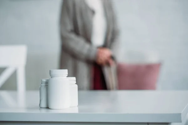 Enfoque selectivo de la mujer mayor con bastón de caminar de pie en la habitación con medicamentos en la mesa en casa - foto de stock