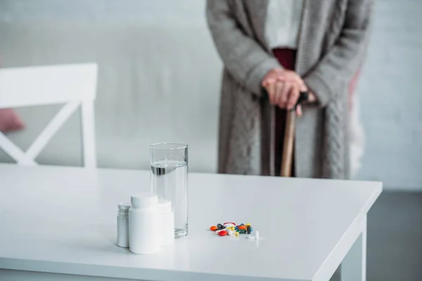 Selective focus of senior woman with walking stick standing in room with medicines and glass of water on tabletop at home — Stock Photo