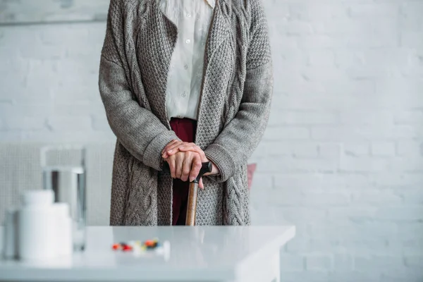 Partial view of senior woman with walking stick standing in room with medicines on tabletop at home — Stock Photo