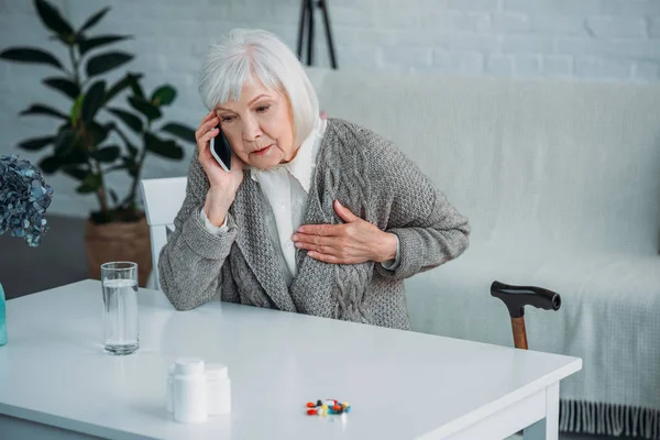 Портрет пожилой женщины с болью в сердце, разговаривающей на смартфоне за столом с лекарствами дома — стоковое фото