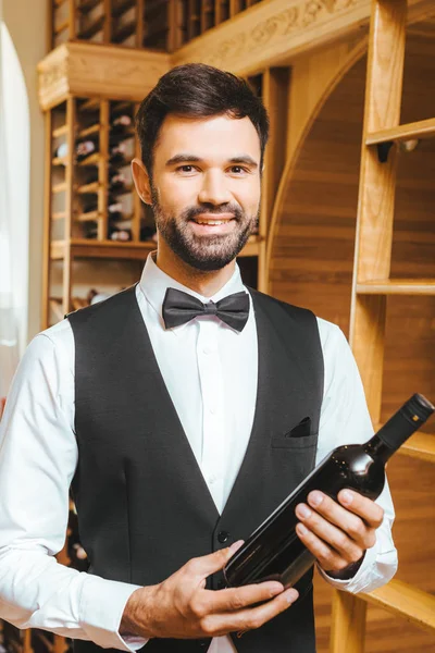 Jovem mordomo sorrindo com garrafa de vinho de luxo na loja de vinhos — Fotografia de Stock