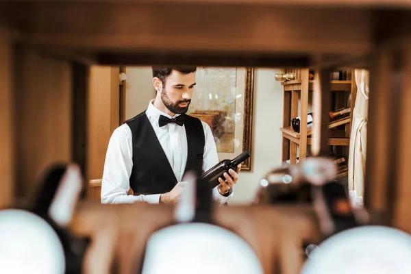 Vue à travers l'étagère à beau jeune steward de vin regardant la bouteille au magasin de vin — Photo de stock