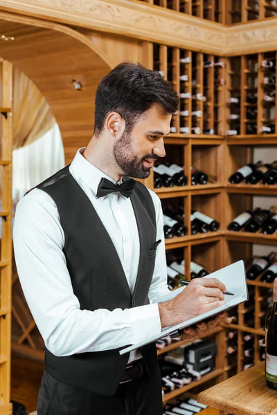 Apuesto joven administrador de vino haciendo notas en la tienda de vinos - foto de stock