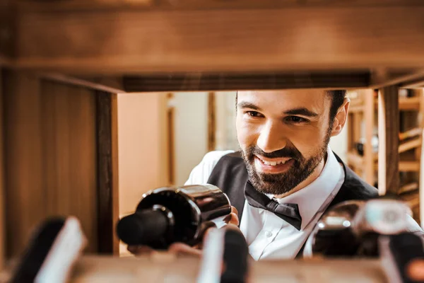 Nahaufnahme eines lächelnden jungen Sommeliers, der eine Flasche im Weinladen ins Regal stellt — Stockfoto