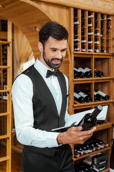 Sonriente joven sommelier mirando la botella de vino y sosteniendo el cuaderno en la tienda de vinos - foto de stock