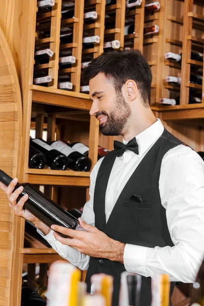 Улыбающийся молодой сомелье смотрит на бутылку вина в винном магазине — стоковое фото