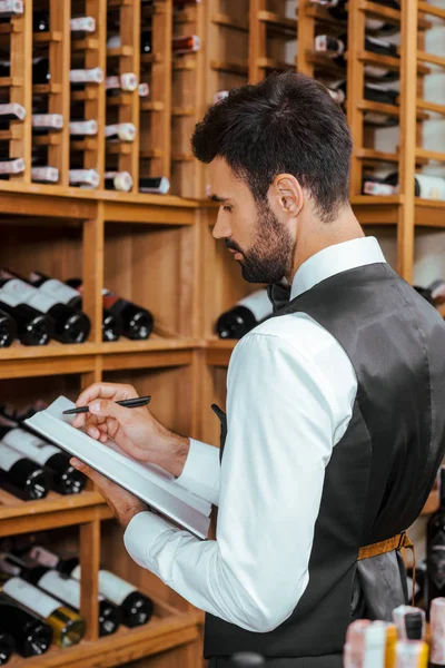 Apuesto joven administrador del vino haciendo notas cerca de estantes en la tienda de vinos - foto de stock