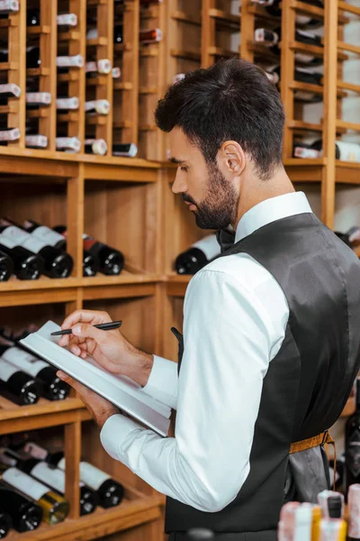 Bello giovane steward prendere appunti vicino scaffali in enoteca — Foto stock