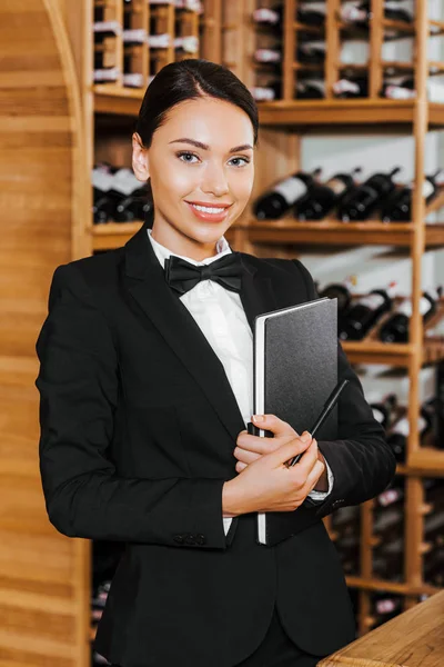 Sonriente mujer administrador de vino witn notebook mirando a la cámara en la tienda de vinos - foto de stock