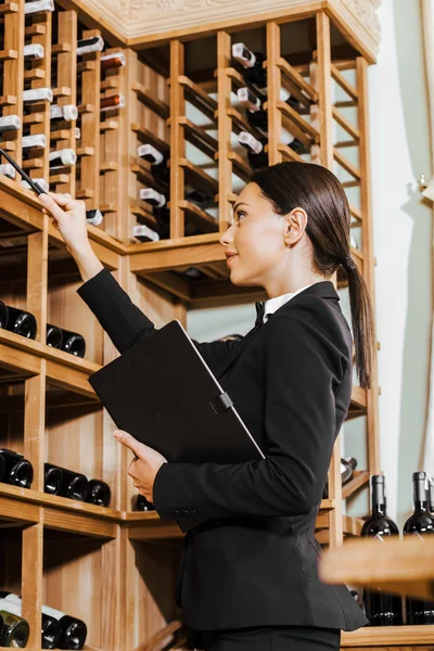 Bela mordomo do vinho feminino com notebook contando garrafas em prateleiras na loja de vinhos — Fotografia de Stock