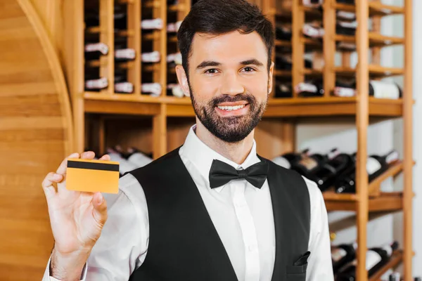 Sonriente joven mayordomo de vino con tarjeta de oro en la tienda de vinos - foto de stock
