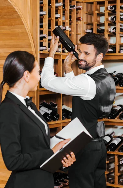 Couple of happy wine stewards doing stocktaking of wine on shelves at store and looking at each other — Stock Photo