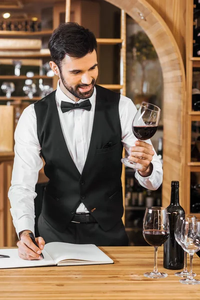 Sonriente joven sommelier examinar el color del vino y tomar notas en la tienda de vinos - foto de stock