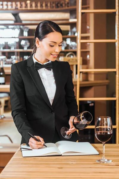 Beautiful female wine steward looking at glass of wine and writing in notebook at wine store — Stock Photo