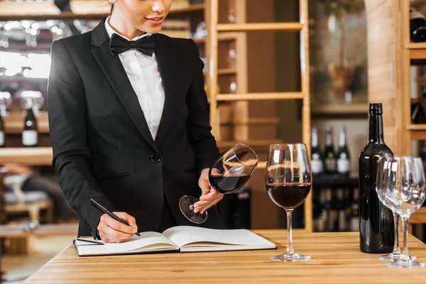 Cropped shot of female wine steward looking at glass of wine and writing in notebook at wine store — Stock Photo