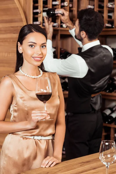 Sonriente mujer sosteniendo vidrio en la tienda de vino mientras mayordomo tomando botella de estante en el fondo - foto de stock