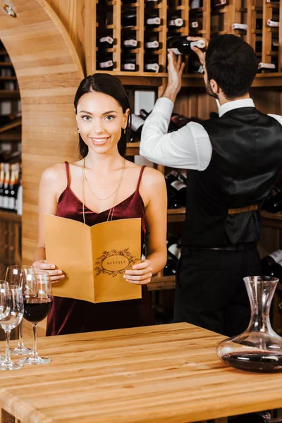Sonriente mujer sosteniendo la tarjeta de menú en la tienda de vinos mientras mayordomo tomando botella de estante en el fondo - foto de stock