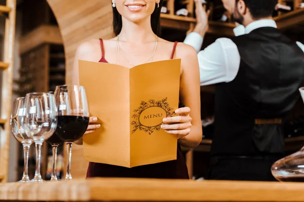 Recortado disparo de la mujer sonriente celebración de la tarjeta de menú en la tienda de vinos mientras mayordomo tomar botella de estante en el fondo - foto de stock