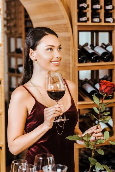 Schöne Frau in rotem Kleid mit Wein und Rose beim Blick auf die Seite im Weinladen — Stockfoto