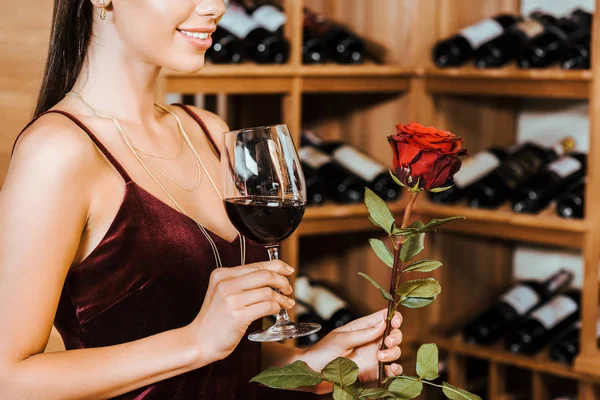 Recortado tiro de hermosa mujer en vestido rojo con vino y rosa en la tienda de vinos - foto de stock