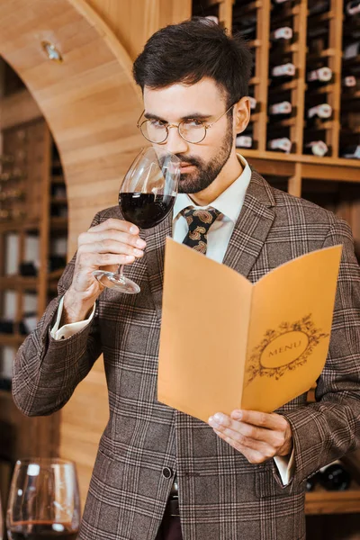Apuesto joven sommelier con carta de menú olfateando vino de vidrio en la tienda de vinos - foto de stock